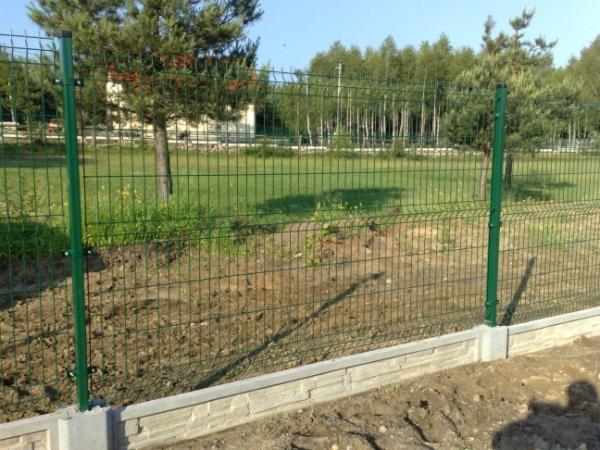 Zielone panele ogrodzeniowe zamontowane na betonowej podmurówce z widocznymi słupkami, w tle trawa i drzewa.