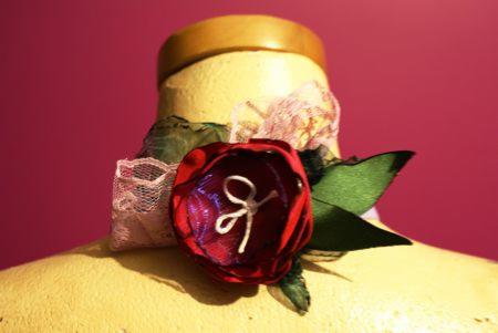 Dekoracyjna opaska z czerwoną, materiałową różą, koronką i zielonymi liśćmi, umieszczona na manekinie o żółtej barwie na tle fuksjowym.