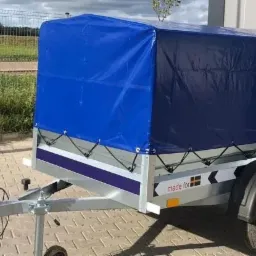 Przyczepa towarowa z niebieską plandeką, widoczne mocowania plandeki, zaczep kulowy i opona, tło stanowi ogrodzenie i budynek.