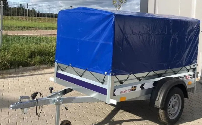 Przyczepa towarowa z niebieską plandeką, widoczne mocowania plandeki, zaczep kulowy i opona, tło stanowi ogrodzenie i budynek.