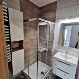 Alex Remonty Paweł Mrozik - Nowoczesna łazienka po remoncie: kabina prysznicowa z chromowanymi profilami, biała szafka pod umywalką z drewnianymi uchwytami, grzejnik drabinkowy i wzorzysta podłoga.