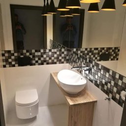 Alex Remonty Paweł Mrozik - Nowoczesna łazienka z wiszącą toaletą, owalną umywalką na drewnianym blacie i dekoracyjną mozaiką nad umywalką, oświetlona przez czarne, stożkowe lampy wiszące.