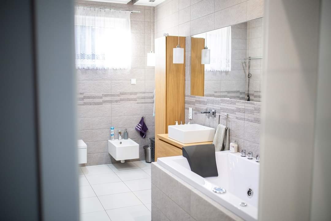 Nowoczesna łazienka z szarymi płytkami, białym bidetem i umywalką, drewnianą szafką, lustrem i wanną z czarnym zagłówkiem, widziana zza framugi drzwi.