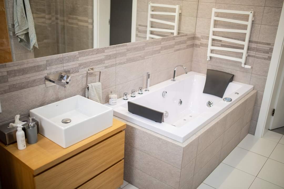 Nowoczesna łazienka z białą wanną z hydromasażem, umywalką nablatową na drewnianej szafce i grzejnikiem drabinkowym, wykończona płytkami w odcieniach szarości.