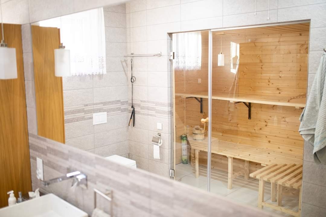 Nowoczesna łazienka z przeszkloną sauną, widoczna umywalka z baterią i toaleta, jasne płytki i drewniane elementy sauny.