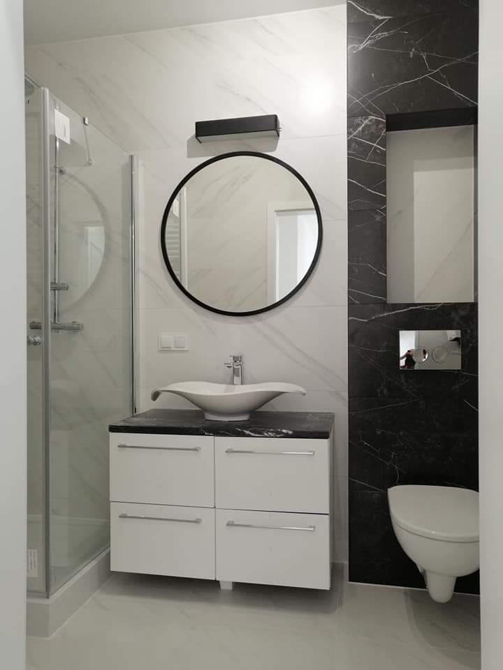 Nowoczesna łazienka z białymi i czarnymi marmurowymi płytkami, okrągłym lustrem i umywalką w kształcie muszli na białej szafce z czarnym blatem, oraz podwieszaną toaletą.
