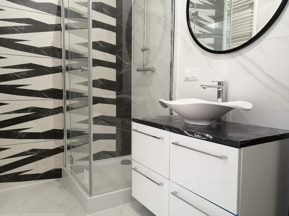 Nowoczesna łazienka z kabiną prysznicową z geometrycznym wzorem na płytkach, białą umywalką w kształcie liścia na szafce z czarnym blatem i okrągłym lustrem.
