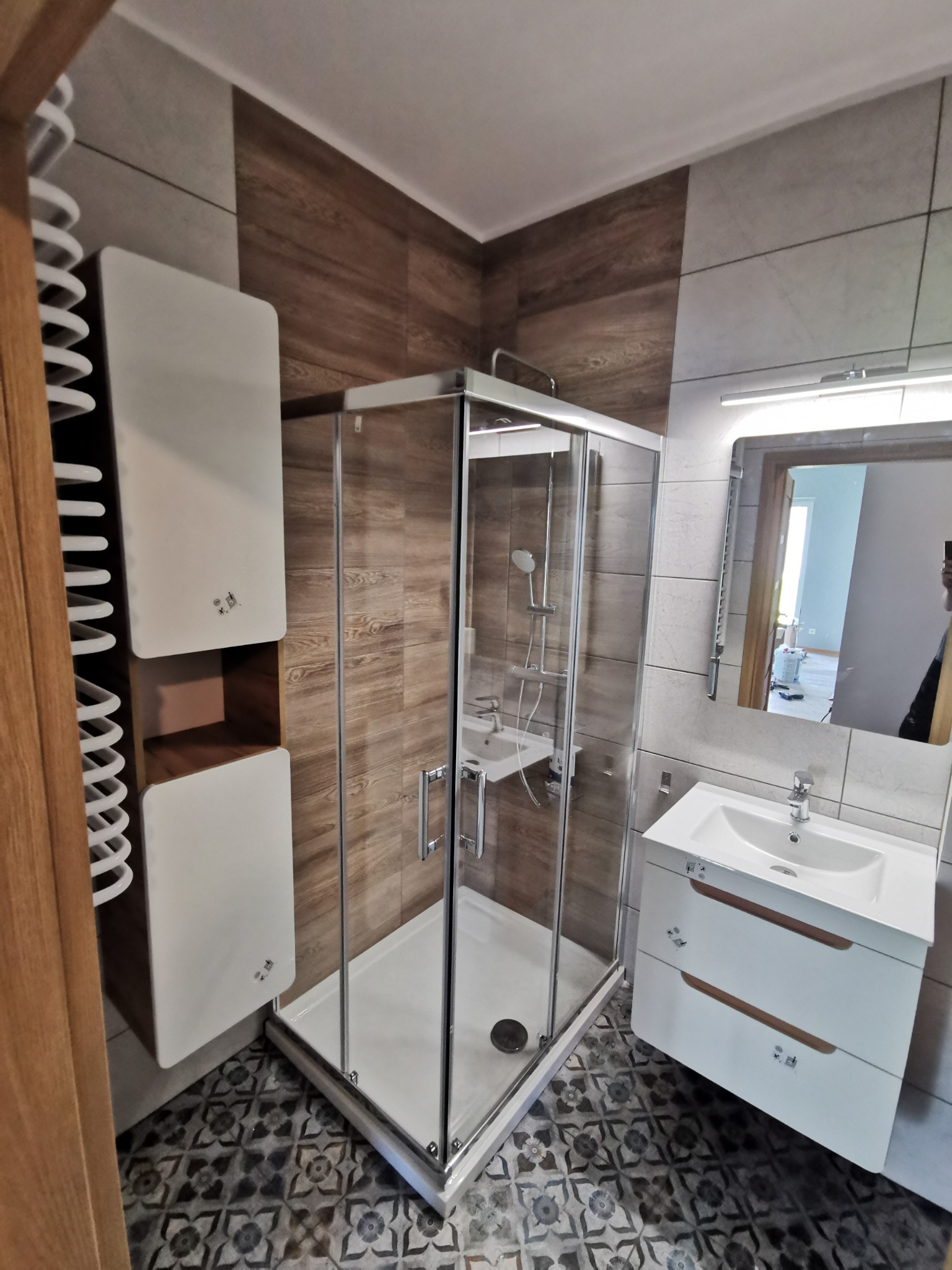 Nowoczesna łazienka po remoncie: kabina prysznicowa z chromowanymi profilami, biała szafka pod umywalką z drewnianymi uchwytami, grzejnik drabinkowy i wzorzysta podłoga.