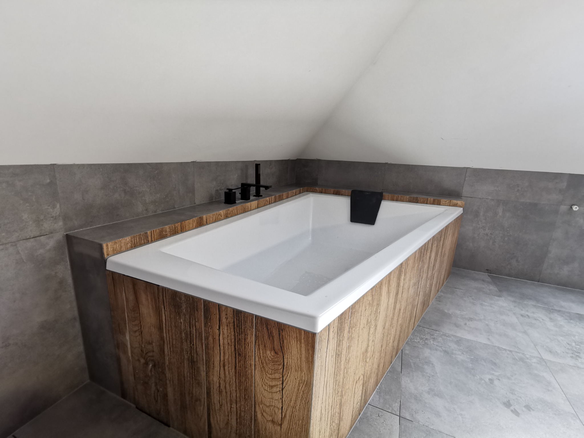 Nowoczesna wanna obudowana panelami imitującymi drewno, z czarną armaturą w łazience wykończonej szarymi płytkami pod skosem dachu.