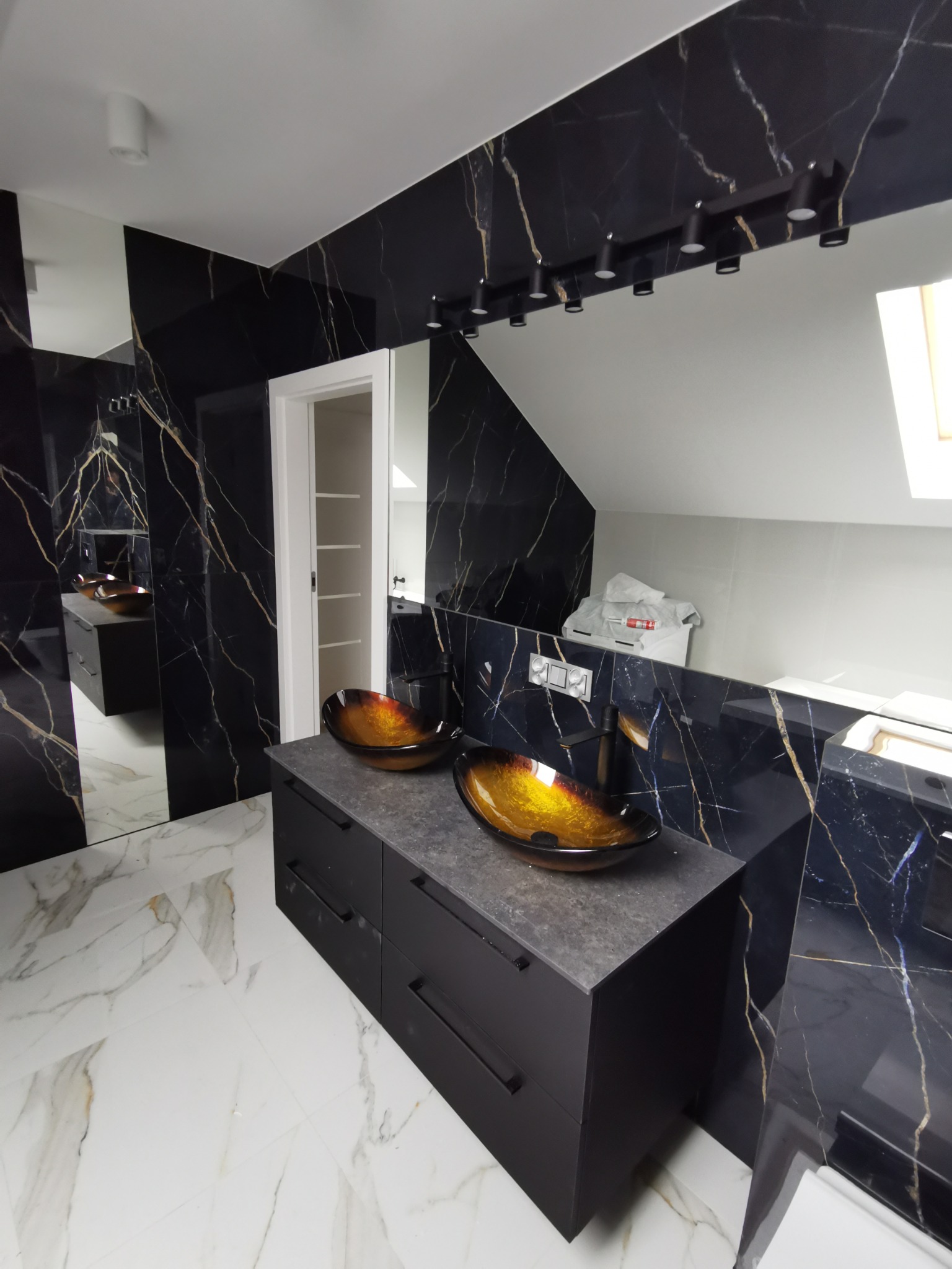 Nowoczesna łazienka z czarnymi płytkami imitującymi marmur ze złotymi żyłami, dwoma szklanymi umywalkami w odcieniach brązu i czarną szafką z szufladami, oświetlenie punktowe na suficie, biała...