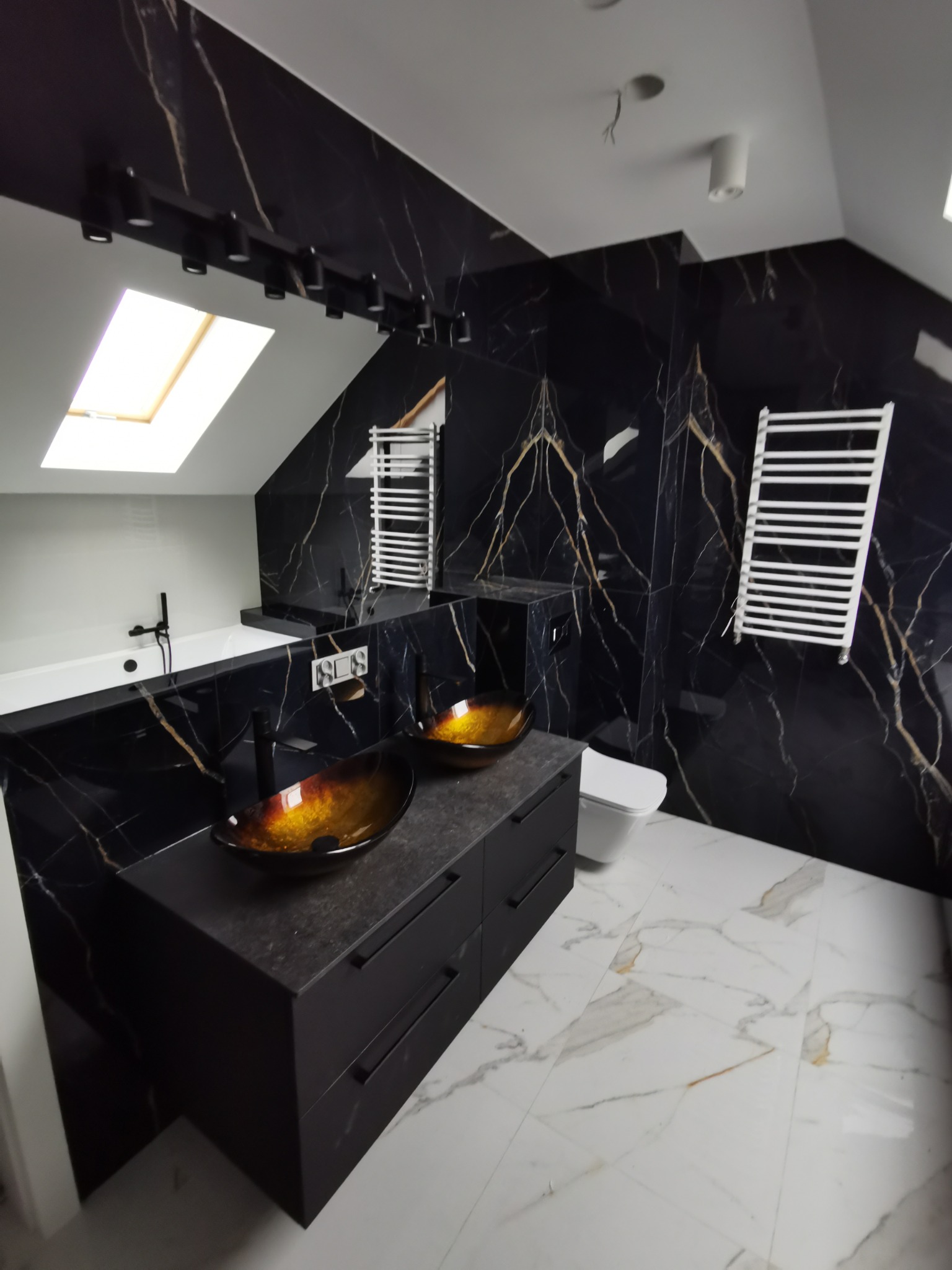 Nowoczesna łazienka na poddaszu z czarnymi płytkami imitującymi marmur ze złotymi żyłami, dwoma szklanymi umywalkami w odcieniach brązu i czarną armaturą, białą wanną i grzejnikami drabinkowymi.