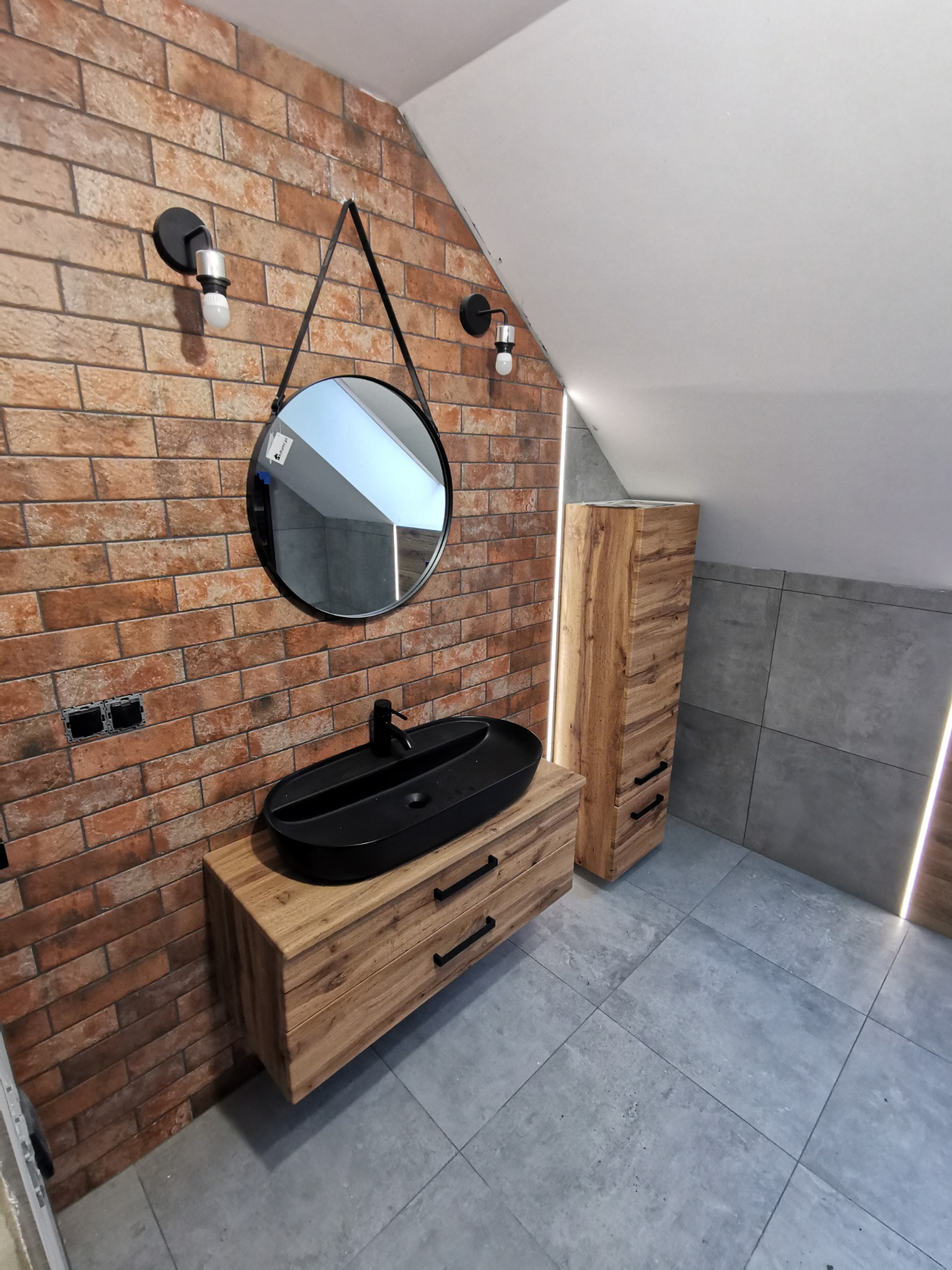 Ukośna łazienka z ceglaną ścianą, okrągłym lustrem na pasku, czarną umywalką na drewnianej szafce i szarą podłogą z płytek, oświetlona listwami LED.
