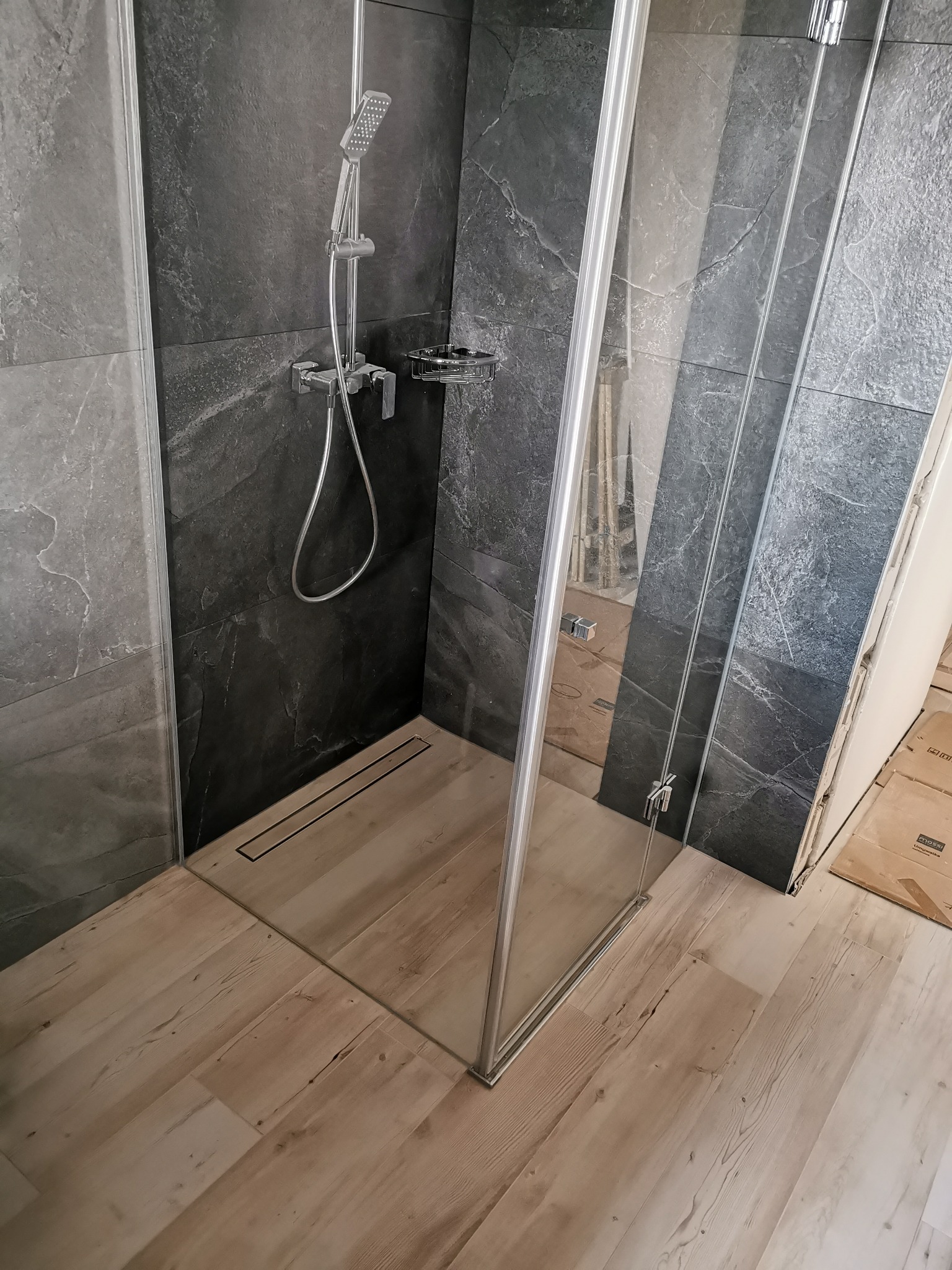Kabina prysznicowa typu walk-in z odpływem liniowym, szare płytki imitujące kamień na ścianach, drewnopodobna podłoga z jasnego drewna.