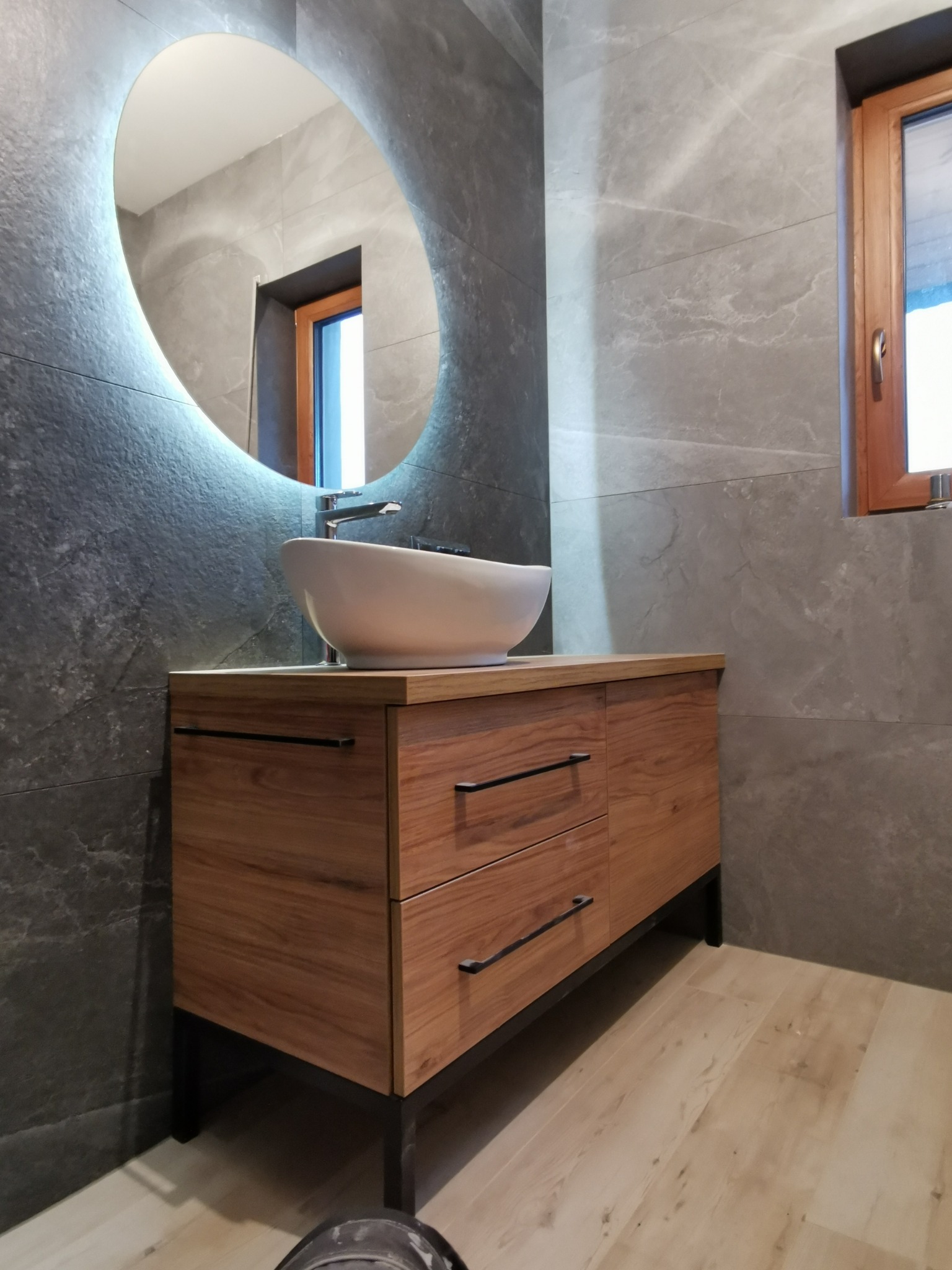 Nowoczesna łazienka z szarą marmurową glazurą, drewnianą szafką pod umywalkę z czarnymi uchwytami i okrągłym lustrem z podświetleniem LED.