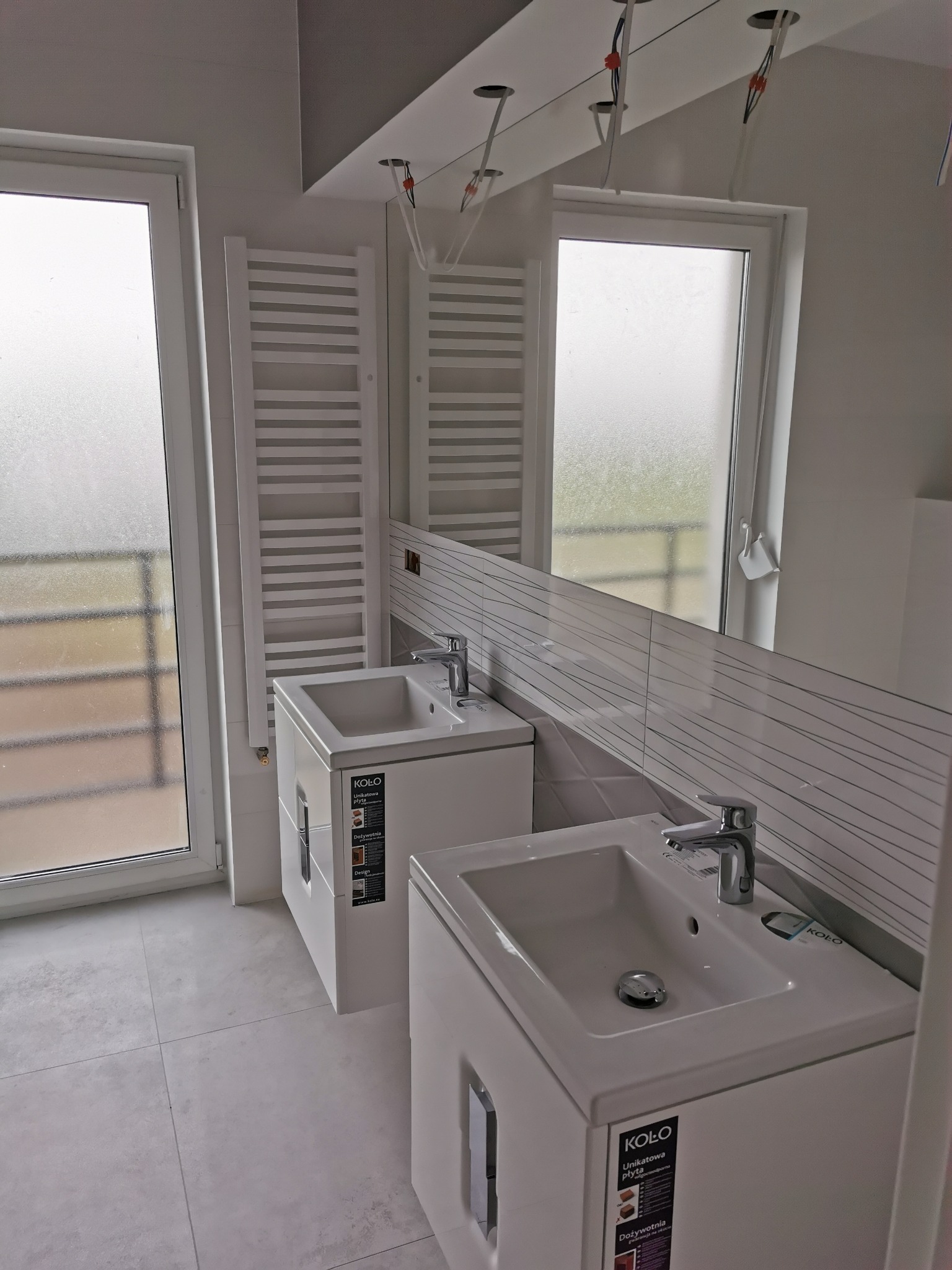 Dwie białe umywalki z szafkami w nowoczesnej łazience z szarymi płytkami, grzejnikiem drabinkowym i dużym lustrem. Widoczne wystające kable oświetleniowe.