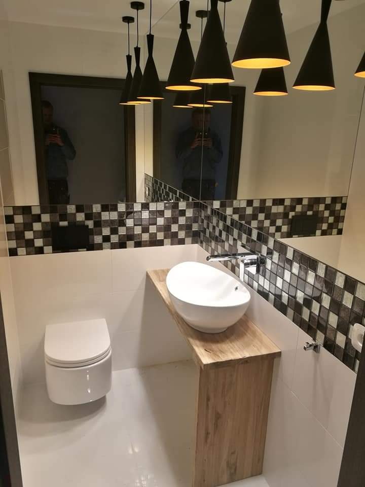 Nowoczesna łazienka z wiszącą toaletą, owalną umywalką na drewnianym blacie i dekoracyjną mozaiką nad umywalką, oświetlona przez czarne, stożkowe lampy wiszące.