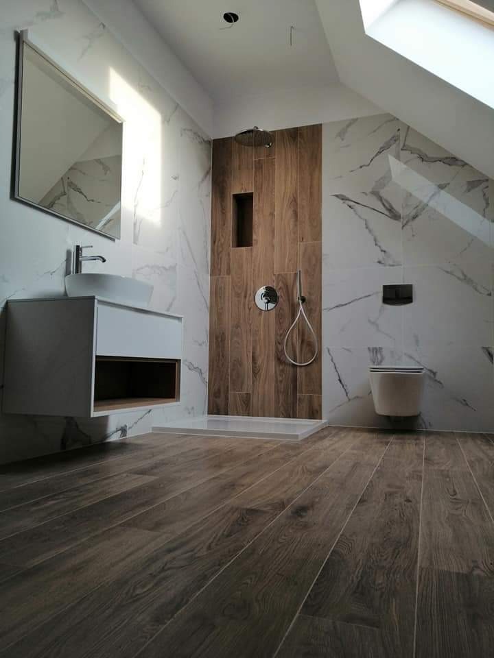 Nowoczesna łazienka na poddaszu z drewnopodobną podłogą i ścianą prysznicową, białe marmurowe płytki na pozostałych ścianach, wisząca umywalka z szafką i lustro.