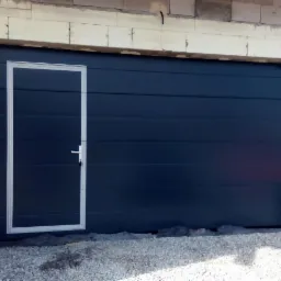 Ciemna brama garażowa segmentowa z drzwiami wejściowymi po lewej stronie, w trakcie montażu, widoczne surowe wykończenie ściany i gruz.