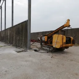 Żółta wiertnica gąsienicowa w trakcie wiercenia otworów w betonowej posadzce pod konstrukcję hali, widoczne betonowe ściany i słupy konstrukcyjne.