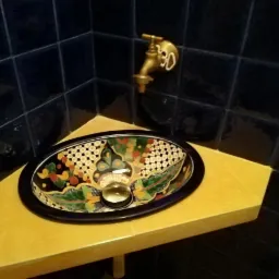 Umywalka ceramiczna w stylu meksykańskim, osadzona na żółtym, trójkątnym blacie w rogu łazienki z ciemnymi, kwadratowymi płytkami i mosiężną armaturą.