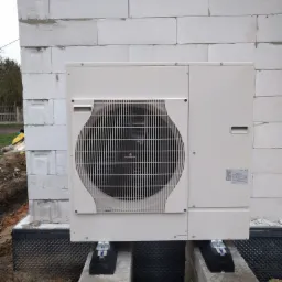 Zewnętrzna jednostka klimatyzacyjna, prawdopodobnie pompy ciepła, zamontowana na betonowych blokach przy ścianie z białego betonu komórkowego. Widoczne elementy montażowe i izolacja fundamentu.