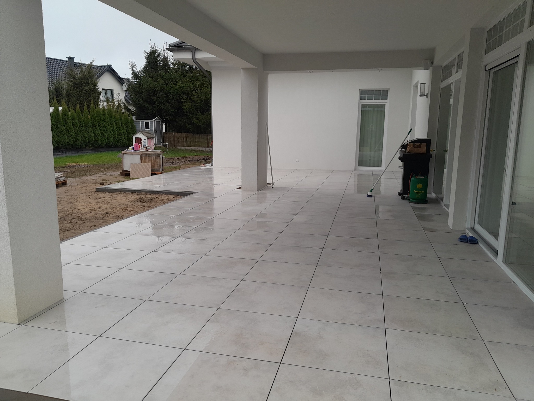 Jasny taras z gresowych płyt, widok zadaszenia, z lewej fragment ogrodu z piaskiem. Na płytkach leżą klapki, stoi grill i butla gazowa.