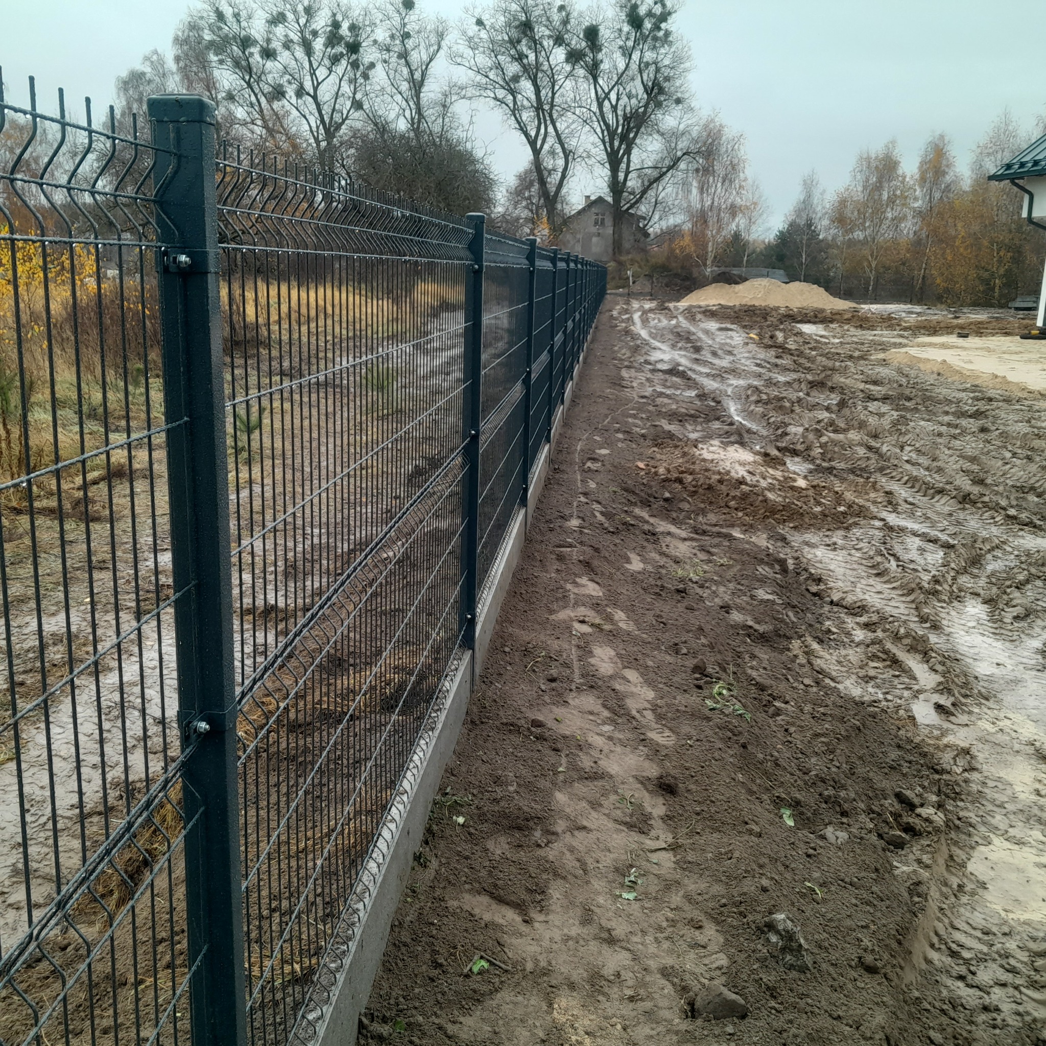 Nowoczesne ogrodzenie panelowe z metalu, z betonową podmurówką, na tle błotnistego terenu i drzew bez liści. Widok z bliska, perspektywa z dołu.