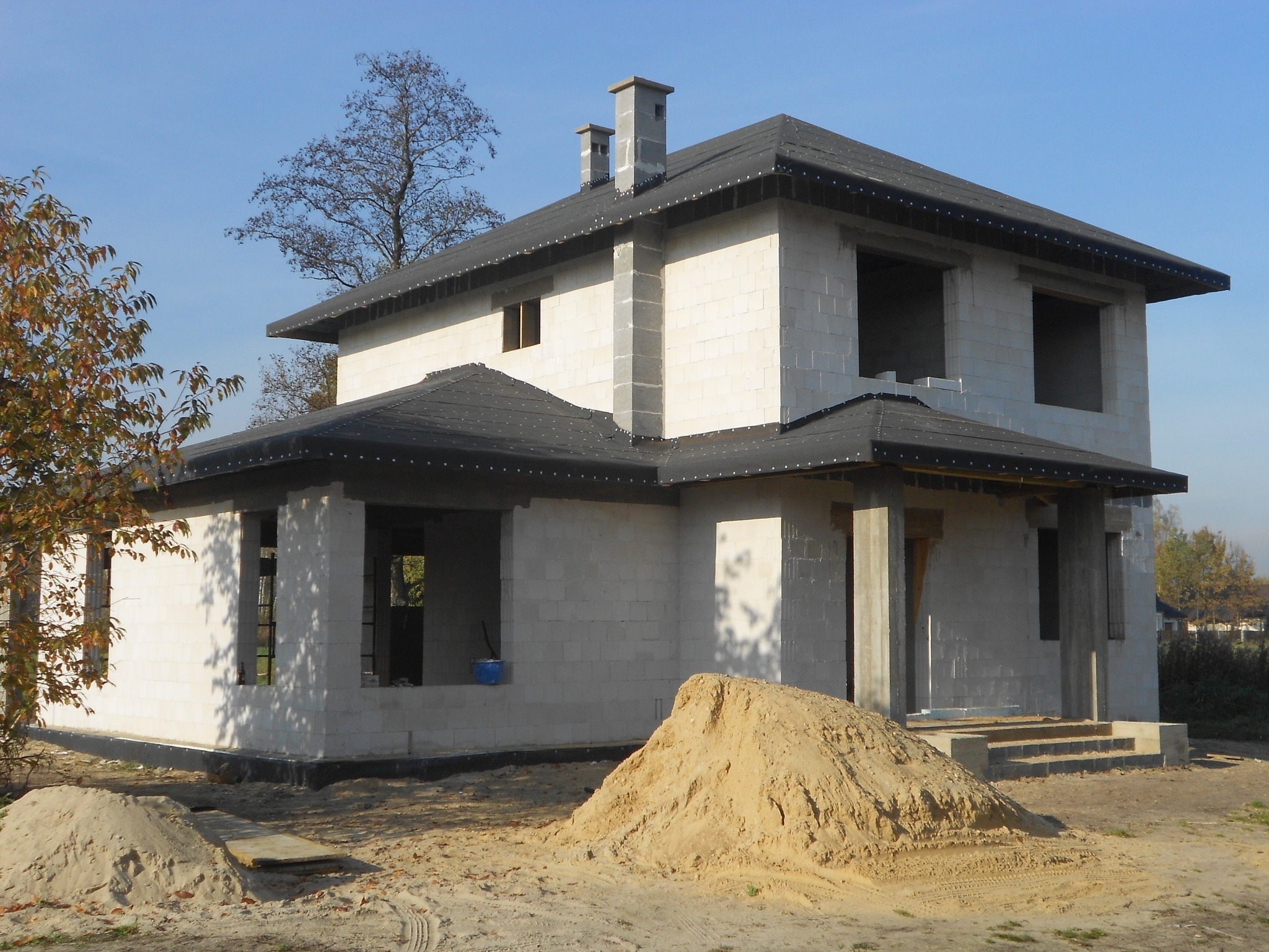 Dwukondygnacyjny budynek w trakcie budowy z białych bloczków, z czarnym dachem i kominami, na działce z piaskiem i drzewami w tle.