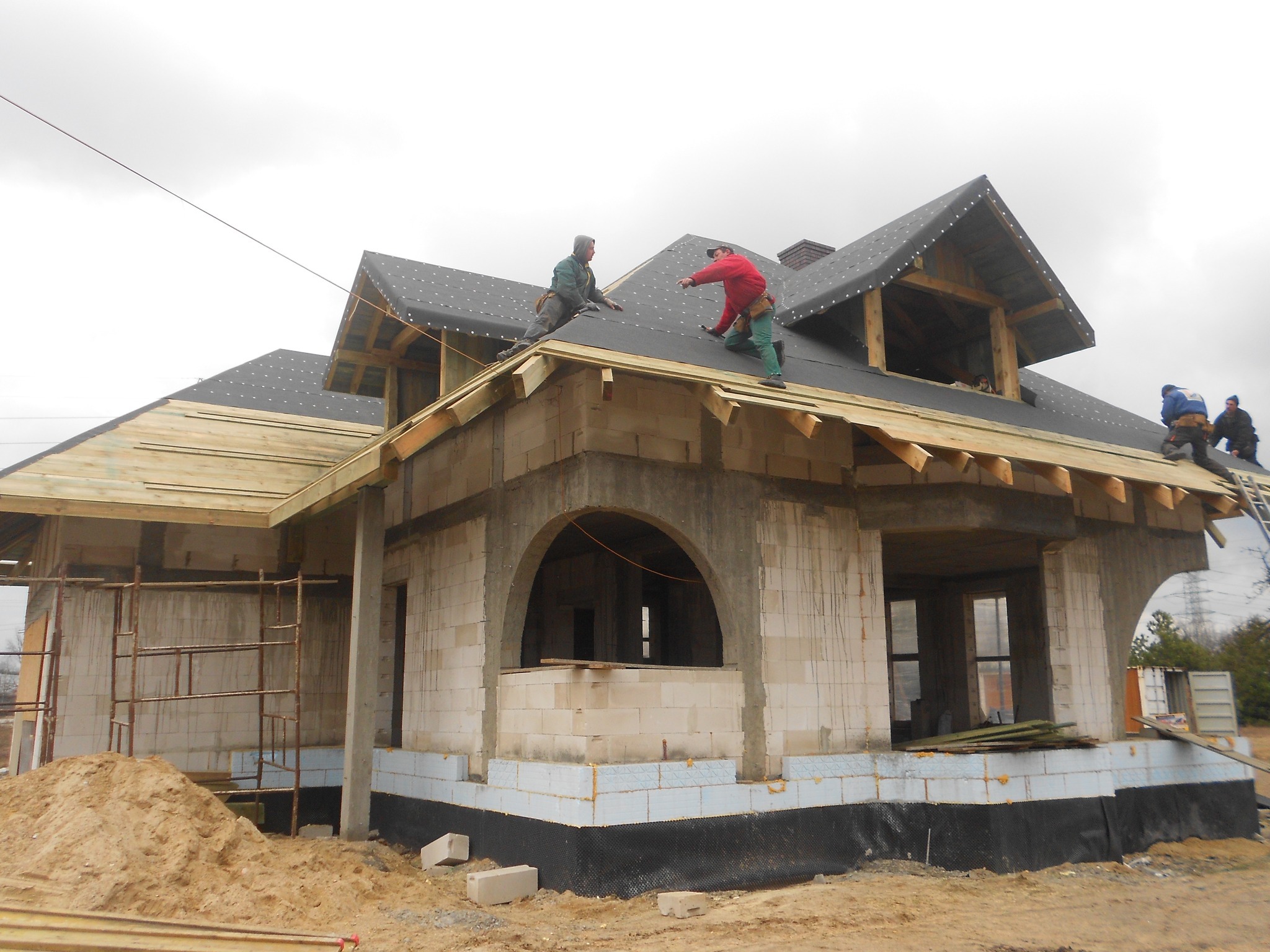 Budowa domu: Murarze pracują na dachu pokrytym papą, widoczne ściany z betonu komórkowego, fundamenty ocieplone styropianem, rusztowania i hałda piasku.