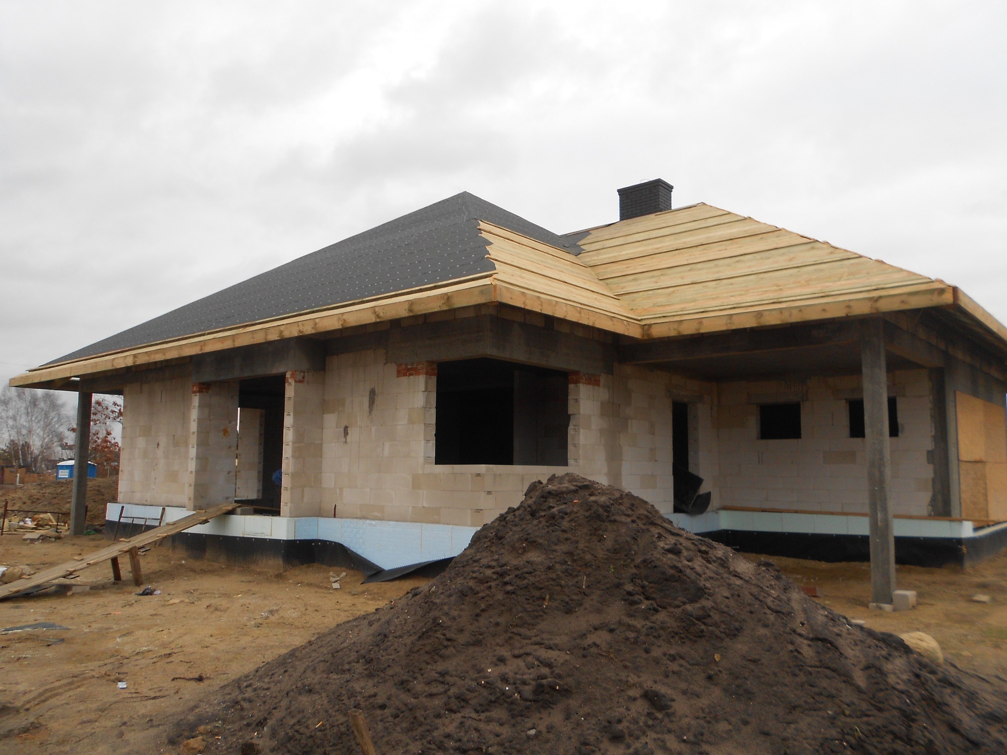 Niedokończony budynek jednorodzinny z pustaków, z częściowo położonym dachem pokrytym papą i deskowaniem, widoczny stos ziemi na pierwszym planie.