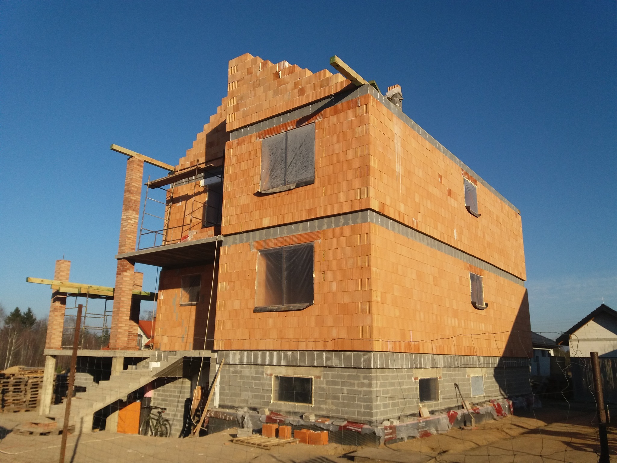 Dwukondygnacyjny dom w trakcie budowy, ściany wzniesione z cegły ceramicznej, okna zabezpieczone folią, widoczne rusztowania i materiały budowlane na placu budowy.