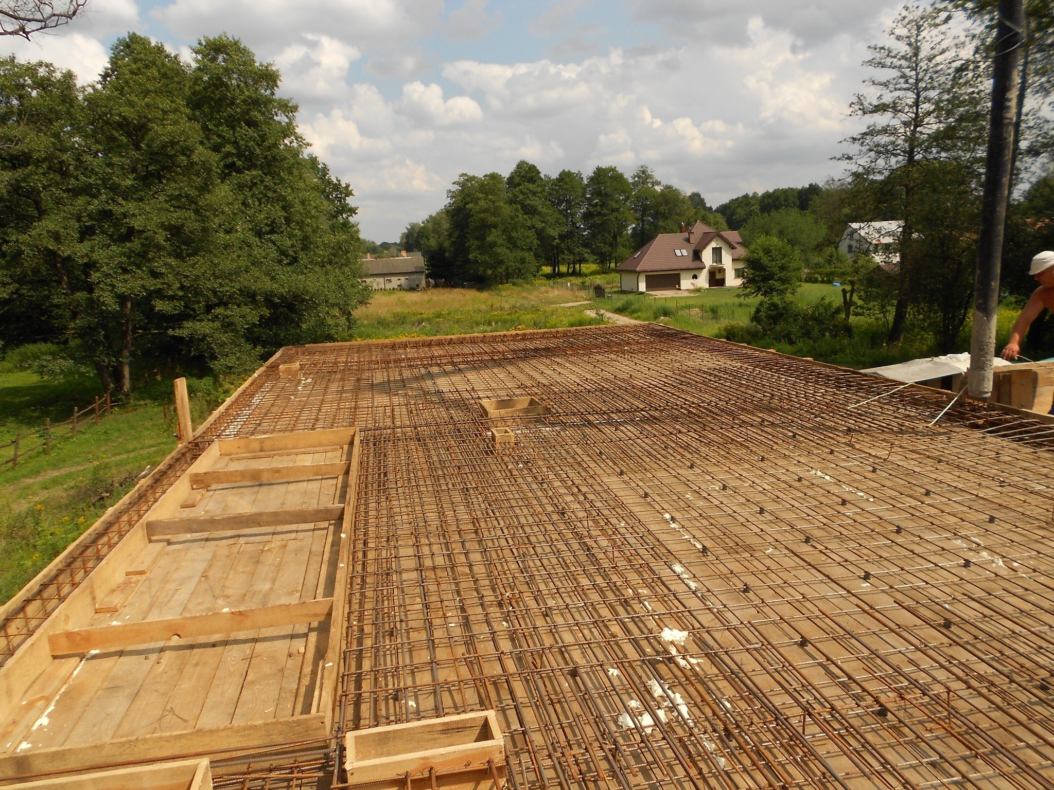 Zbrojenie stropu na deskowaniu, widok z góry na siatkę z prętów stalowych i drewniane szalunki, w tle zabudowa jednorodzinna i zieleń