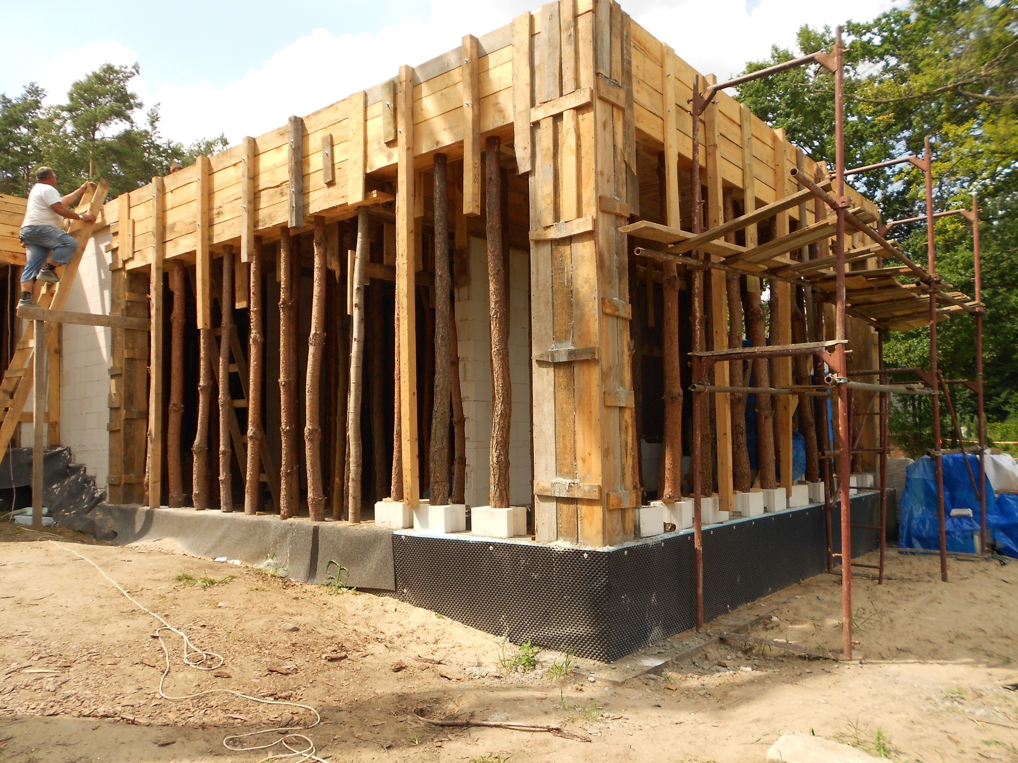 Budowa domu murowanego: szalunek z drewnianych desek i okrąglaków, widoczny fundament z izolacją, pracownik na drabinie montujący elementy konstrukcji.