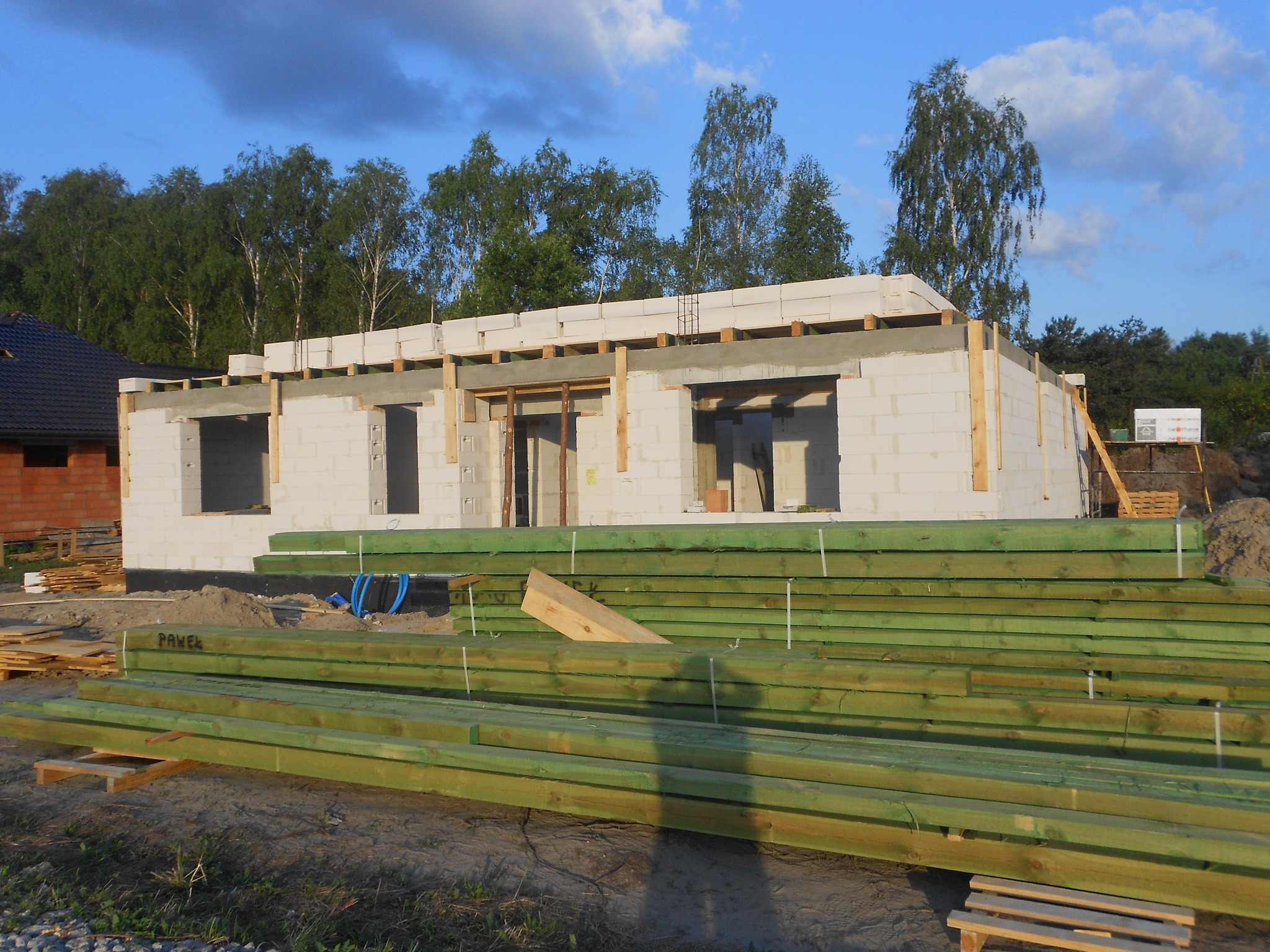Surowy stan domu z białego bloczka, w tle drzewa i błękitne niebo z chmurami, na pierwszym planie stos zielonych impregnowanych desek z napisem 'PAWEŁ'.