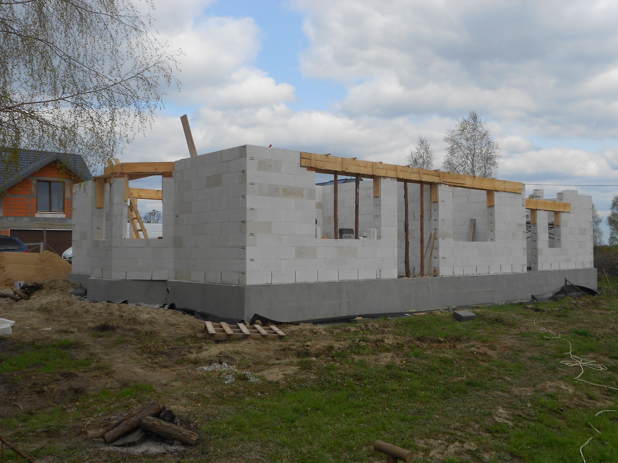 Wznoszenie ścian parterowego domu z betonu komórkowego, widoczne drewniane nadproża okienne, w tle dom z czerwonej cegły.