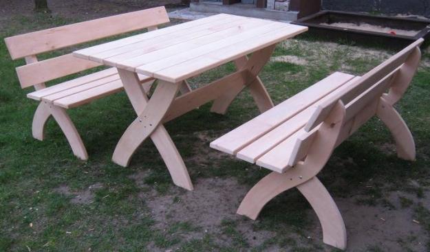 Zestaw ogrodowy z jasnego drewna: stół i dwie ławki z oparciami, o charakterystycznych nogach w kształcie litery X, ustawiony na trawniku.