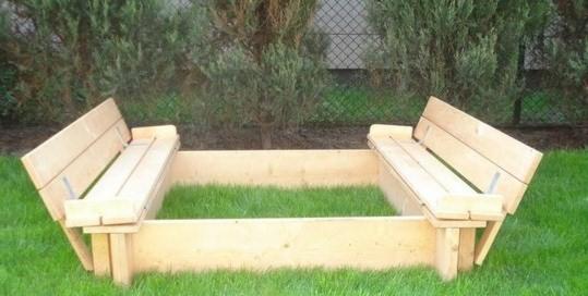 Drewniana piaskownica z wbudowanymi ławeczkami, wypełniona trawą, na tle trawnika i żywopłotu.