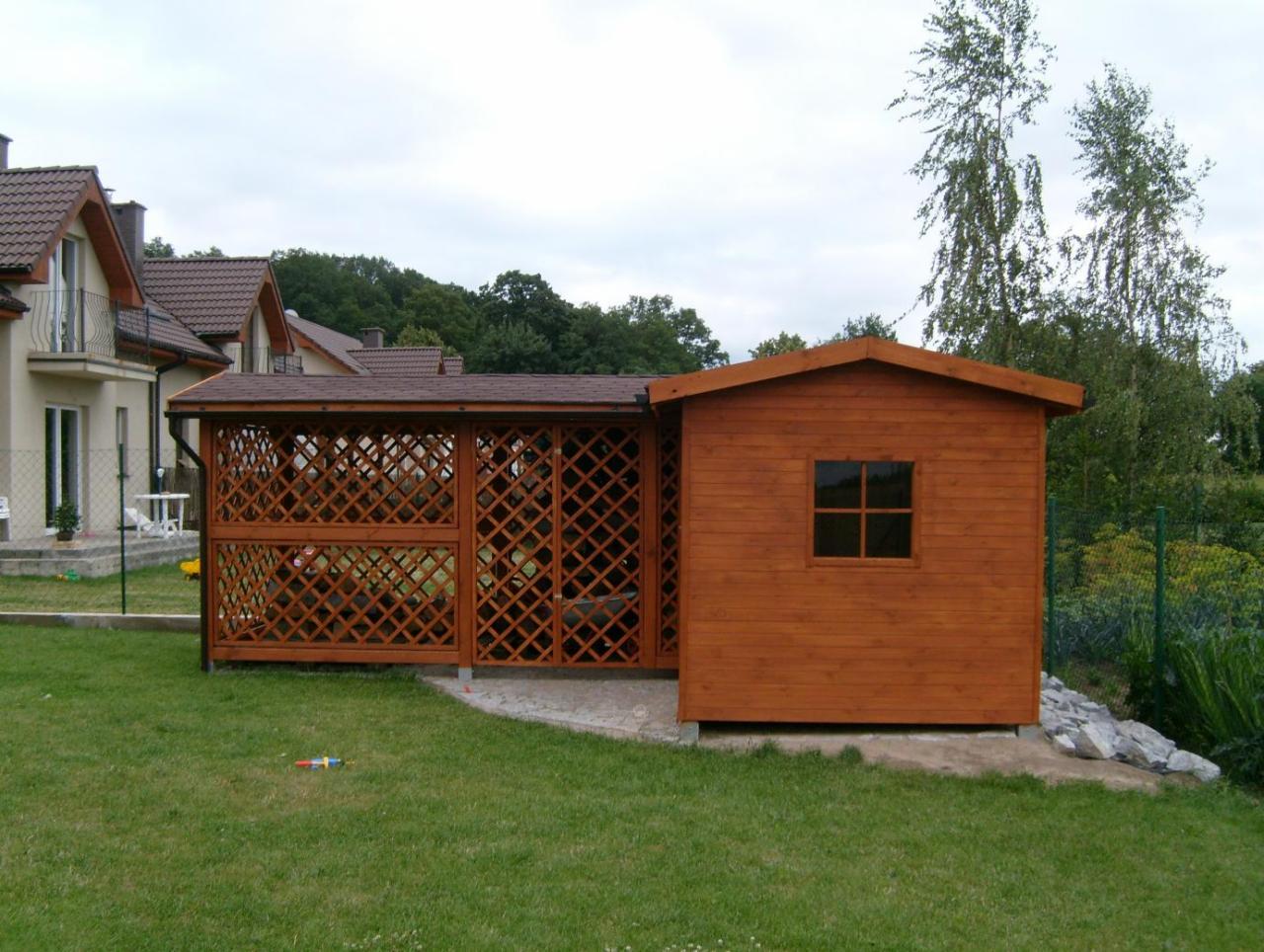 Drewniana altana z ażurową ścianą i małym domkiem, brązowy dach, zielony trawnik, domy w tle.