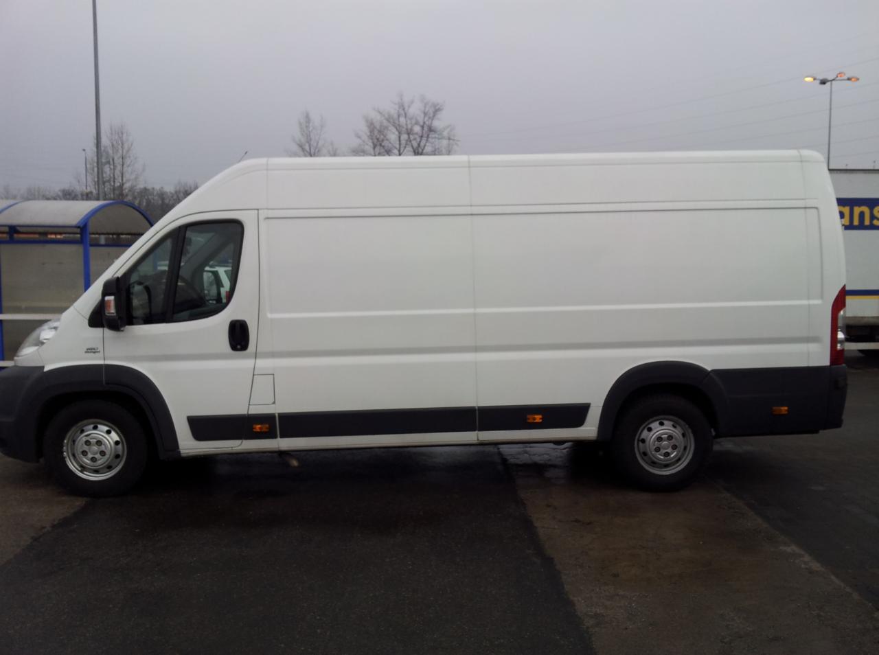 Biały, dostawczy bus z wysokim dachem, zaparkowany na mokrym asfalcie w pochmurny dzień. Widok z boku, zarys drzew i wiaty przystankowej w tle.