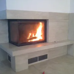 Nowoczesny kominek obudowany jasnym trawertynem, z widocznym płomieniem ognia. Minimalistyczny design z prostymi liniami i dyskretnymi otworami wentylacyjnymi.