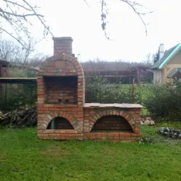 Murowany grill ogrodowy z czerwonej cegły, z paleniskiem i miejscem na drewno, na tle zielonego trawnika i budynku z zielonym dachem.