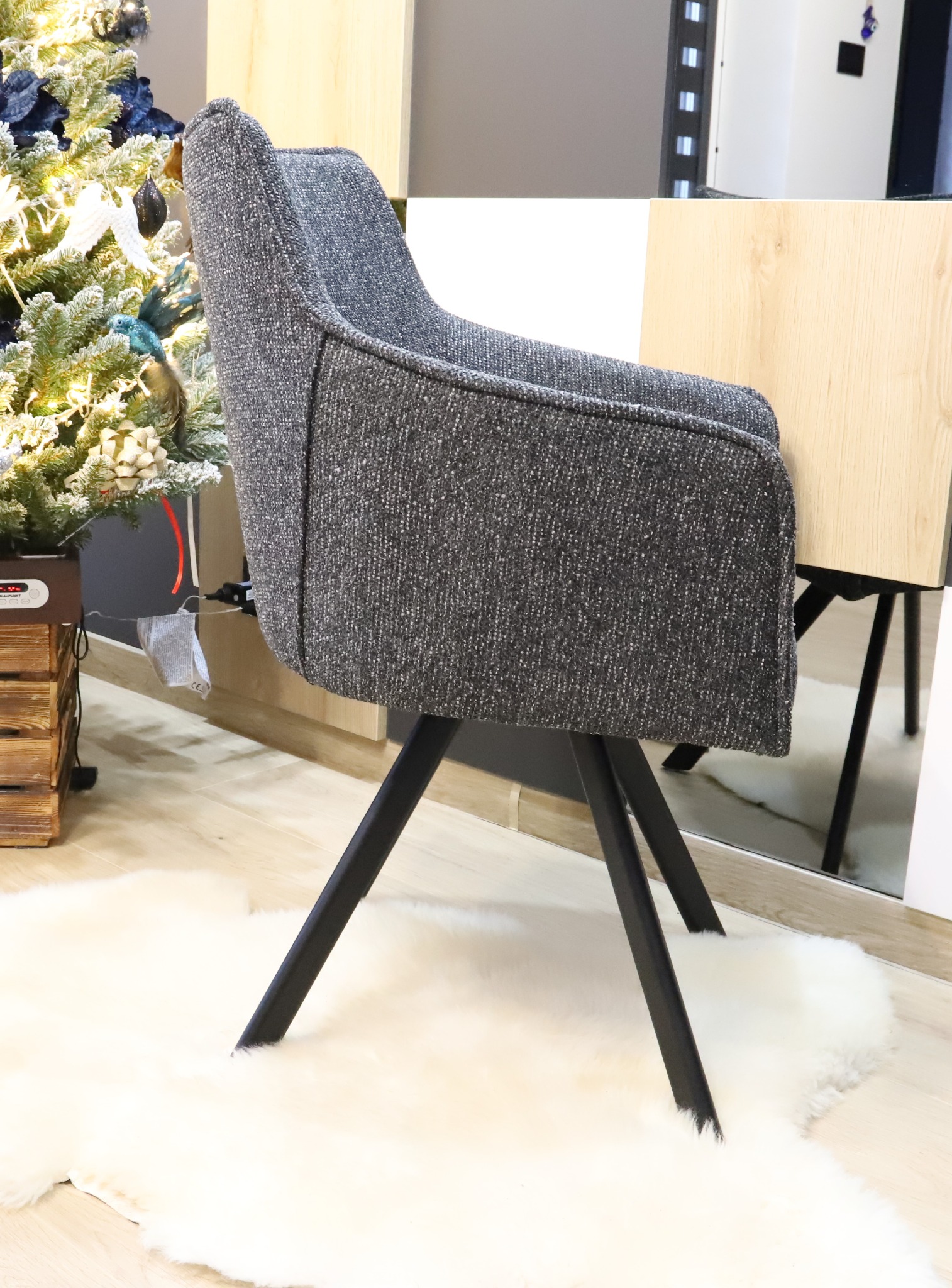 Tapicerowane krzesło w odcieniach szarości na czarnych, skośnych nogach, stoi na białym dywanie z długim włosiem, obok choinki z niebieskimi ozdobami.