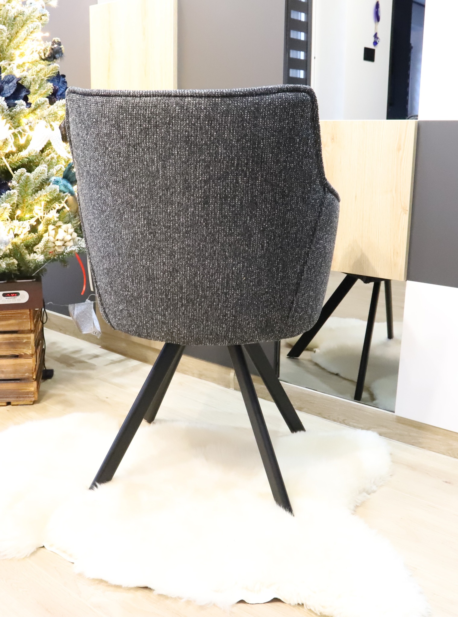 Szare krzesło tapicerowane z ciemnymi nogami na białym dywanie z długim włosiem, obok choinki i szafki z elementami drewna i szarości.