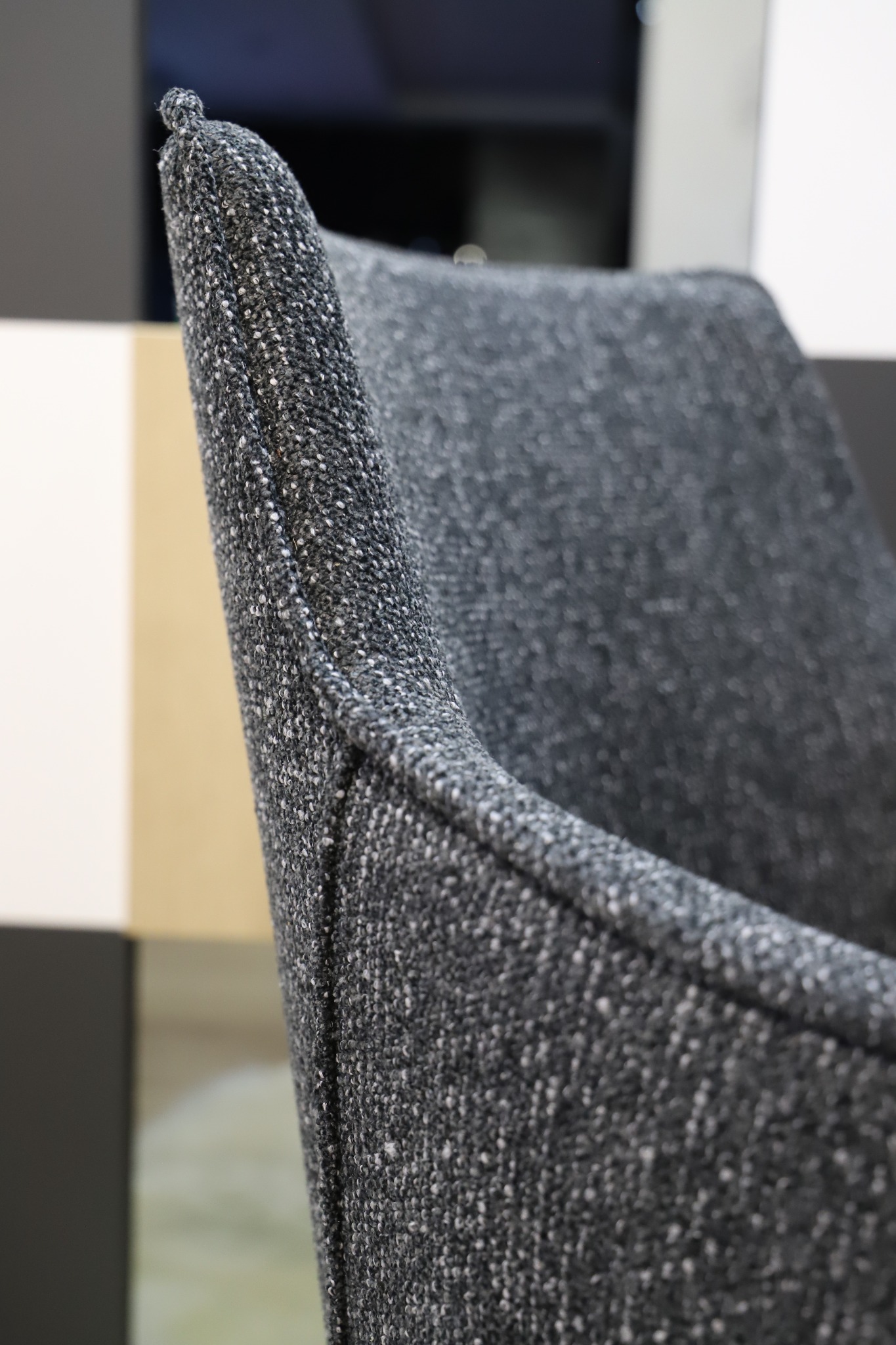 Fragment szarego krzesła tapicerowanego tkaniną melanżową, z widocznym szwem i zaokrąglonym wykończeniem oparcia.