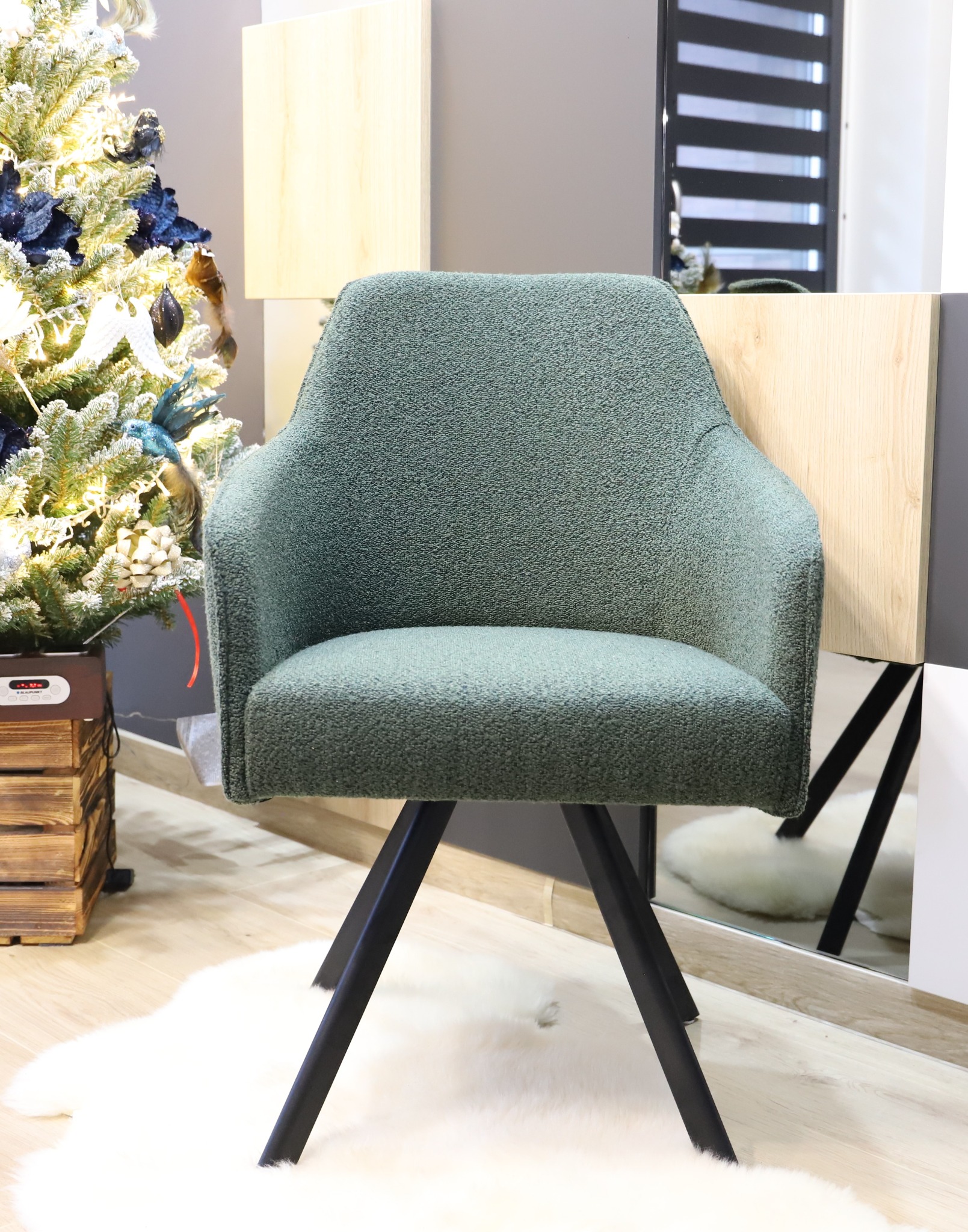 Turkusowe krzesło tapicerowane na czarnych nogach, ustawione na białym dywanie z długim włosiem, obok choinki z niebieskimi ozdobami.