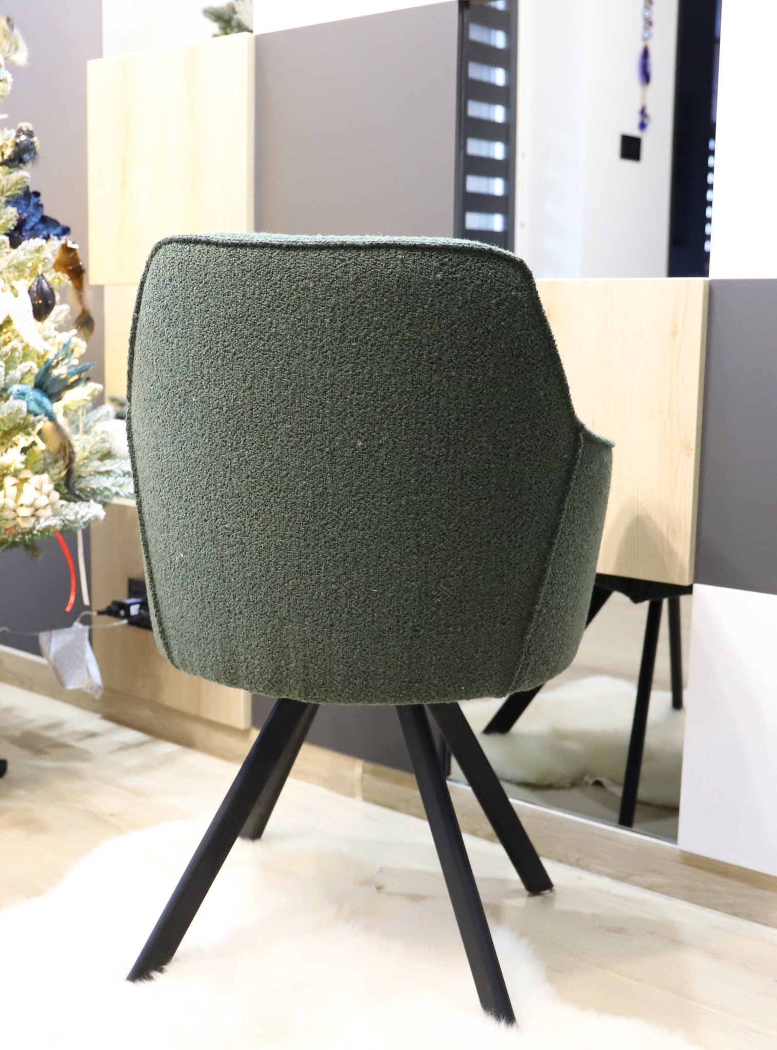 Krzesło tapicerowane w odcieniach zieleni, ustawione na białym, puszystym dywanie, z widocznym fragmentem świątecznego drzewka w tle.