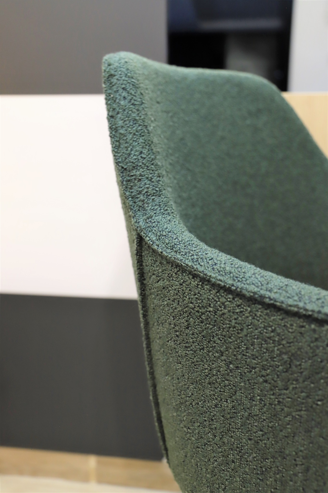 Zbliżenie na krawędź tapicerowanego krzesła w odcieniu zieleni, z widoczną fakturą materiału na tle ściany w kolorach bieli i szarości.