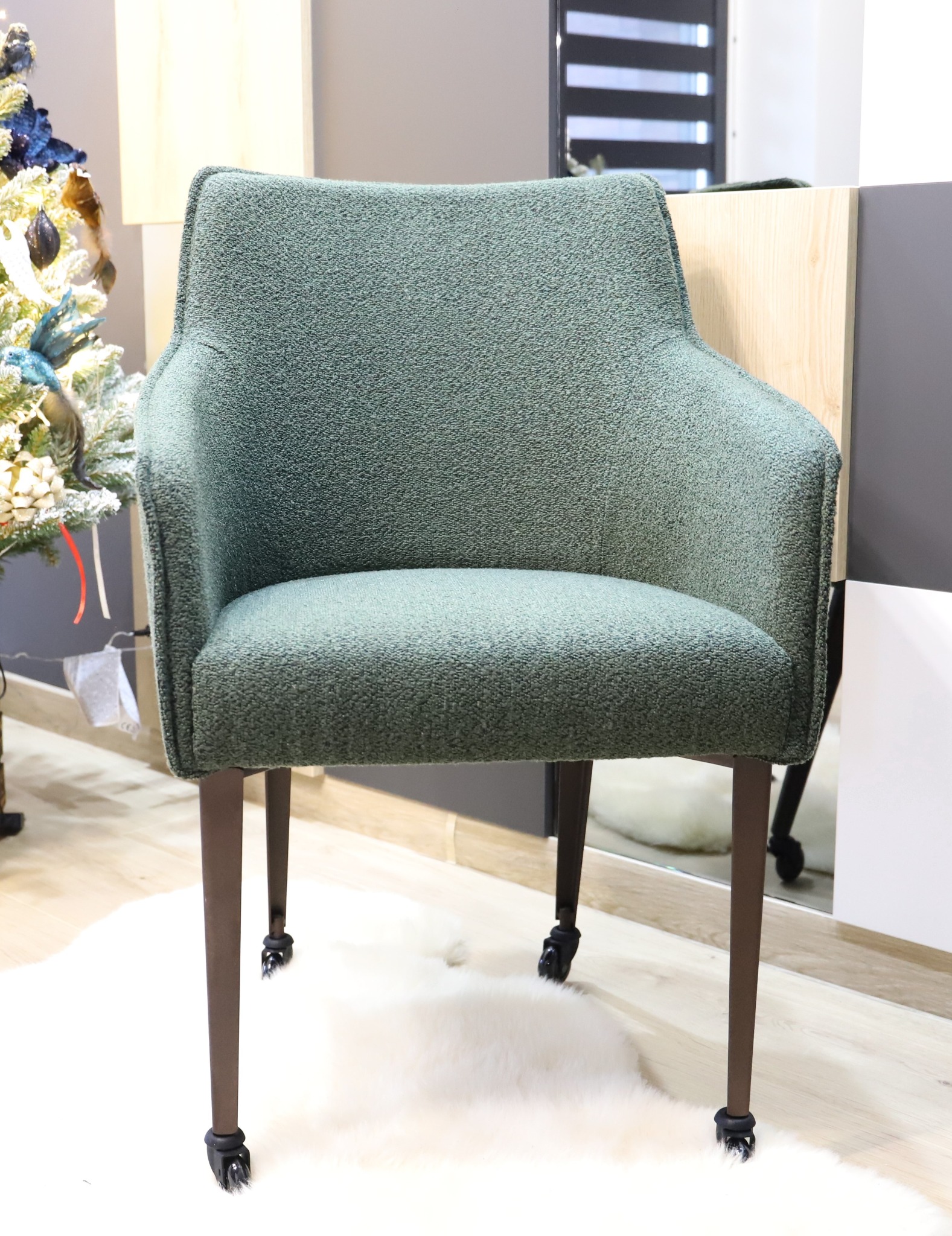 Zielone krzesło tapicerowane z podłokietnikami i brązowymi nogami na kółkach, stojące na białym futrzanym dywanie, obok choinki i nowoczesnej komody.