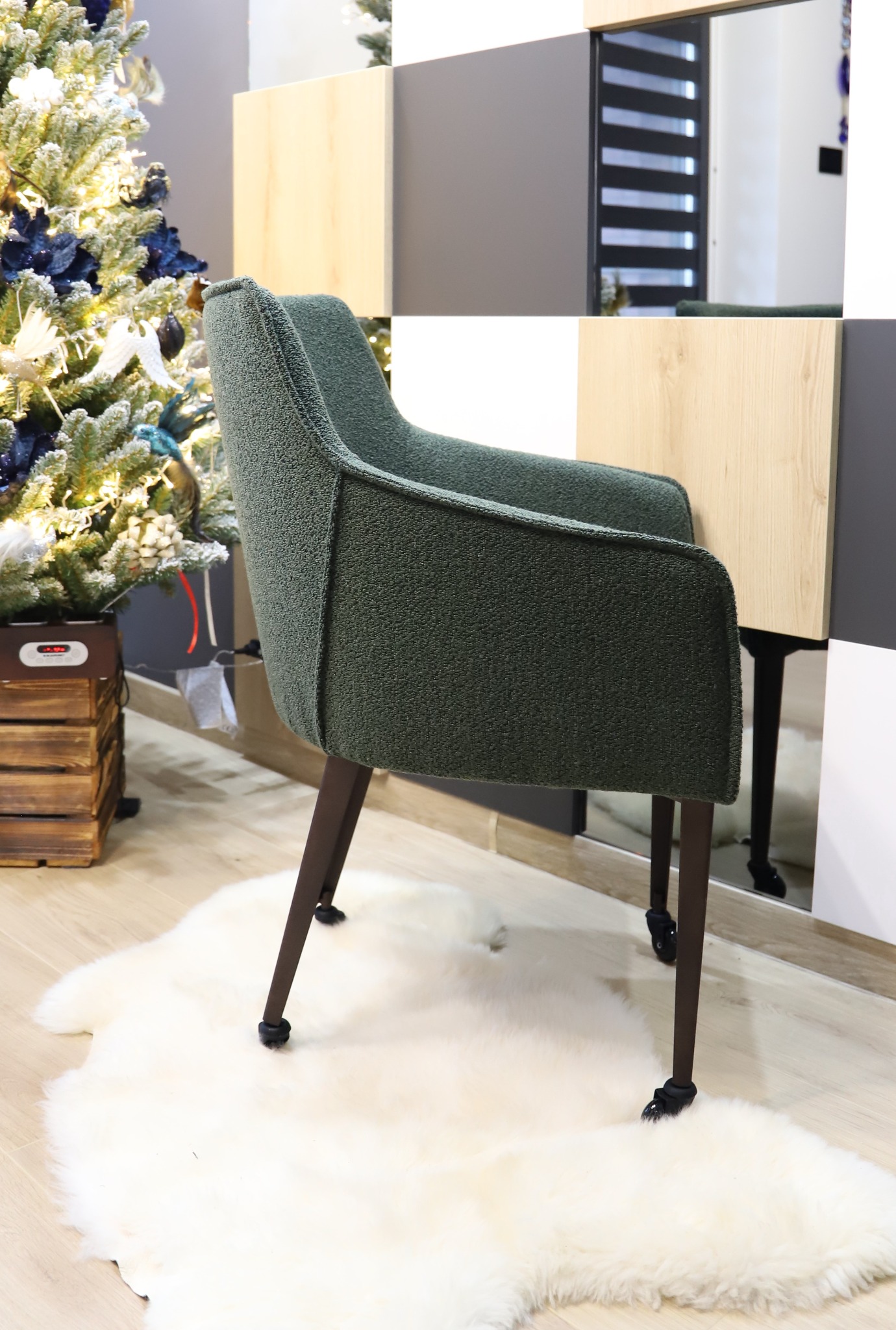 Zielony fotel tapicerowany z podłokietnikami i brązowymi nogami na kółkach, stojący na białym dywanie ze sztucznego futra, obok choinki i zabudowy meblowej z elementami drewnopodobnymi.