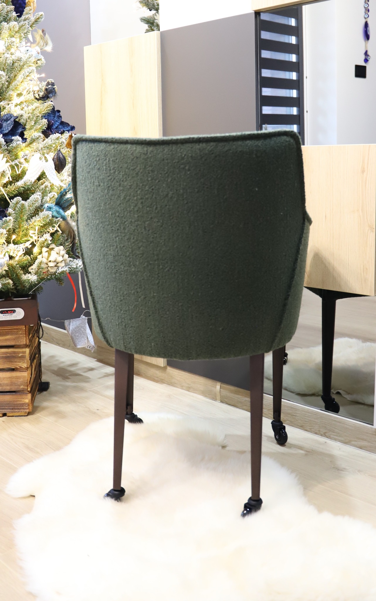 Zielone krzesło tapicerowane z brązowymi nogami na kółkach, widziane od tyłu, na tle jasnej podłogi, białego dywanu z długim włosiem, choinki i szafki z lustrem.